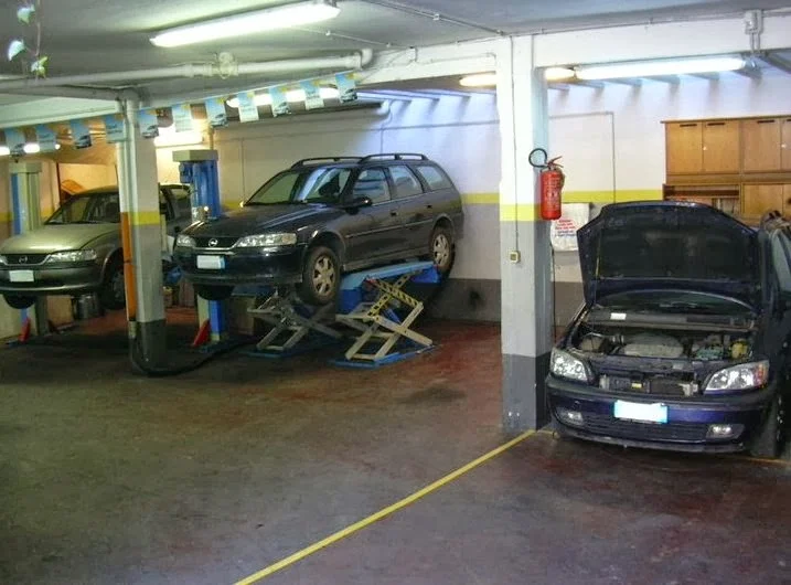 L'interno dell'officina: due auto sui ponti sollevatori, un'altra con il cofano aperto, pareti chiare con le fasce gialle